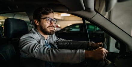 Homem jovem de barba e óculos no interior de um carro estacionado numa garagem.