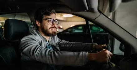 Homem jovem de barba e óculos no interior de um carro estacionado numa garagem.