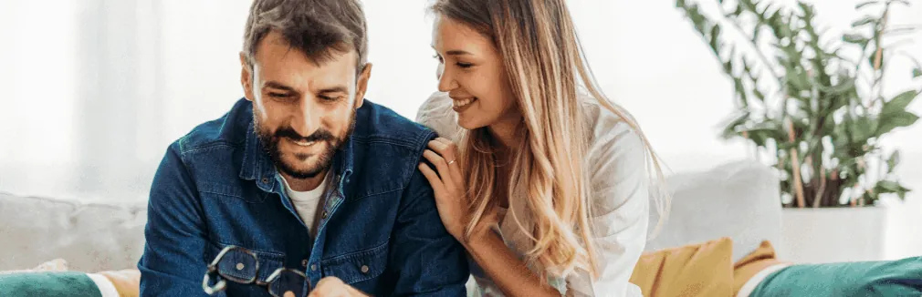 Casal jovem, sentado num sofá, a olhar para um tablet e a sorrir 