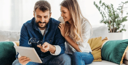 Casal jovem, sentado num sofá, a olhar para um tablet e a sorrir 