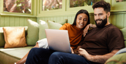 Casal sorridente faz um seguro de vida online, sentado no sofá com portátil nos joelhos