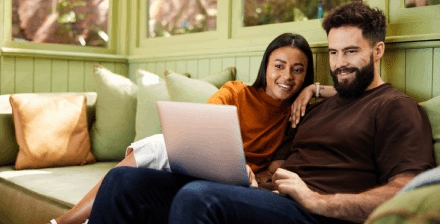 Casal sorridente sentado no sofa verifica portatil Casal sorridente faz um seguro de vida online, sentado no sofá com portátil nos joelhos